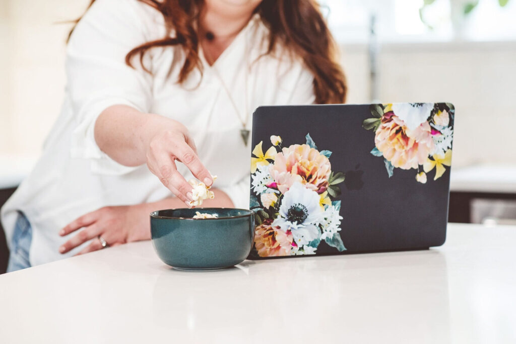 Woman reaching for a bowl of popcorn while working at a floral-covered laptop, representing a moment of emotional eating at home.

