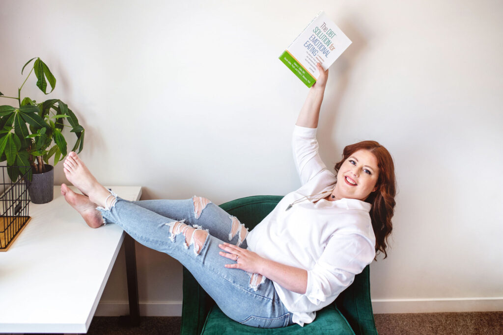 Woman smiling while lounging in a chair, holding book about how to stop emotional eating.