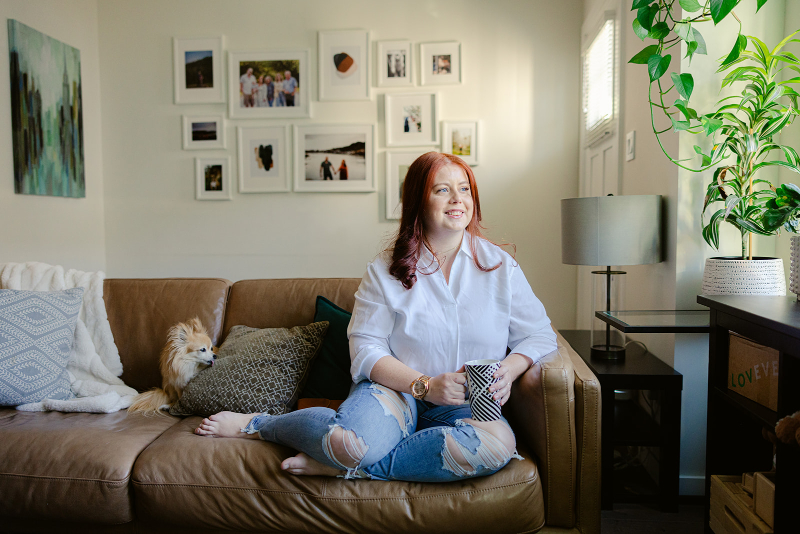 Caucasian female therapist sitting on a brown leather sofa, while holding a coffee cup. She's looking out the window while smiling. Her pomeranian-chihuahua dog sits beside her.