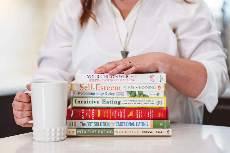 a person in a white shirt sitting at a table, holding a white textured mug in one hand and resting the other hand on a stack of books.