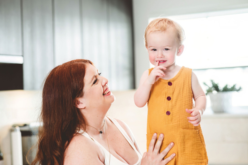 Woman smiling at toddler in yellow romper, highlighting connection while navigating food talk.