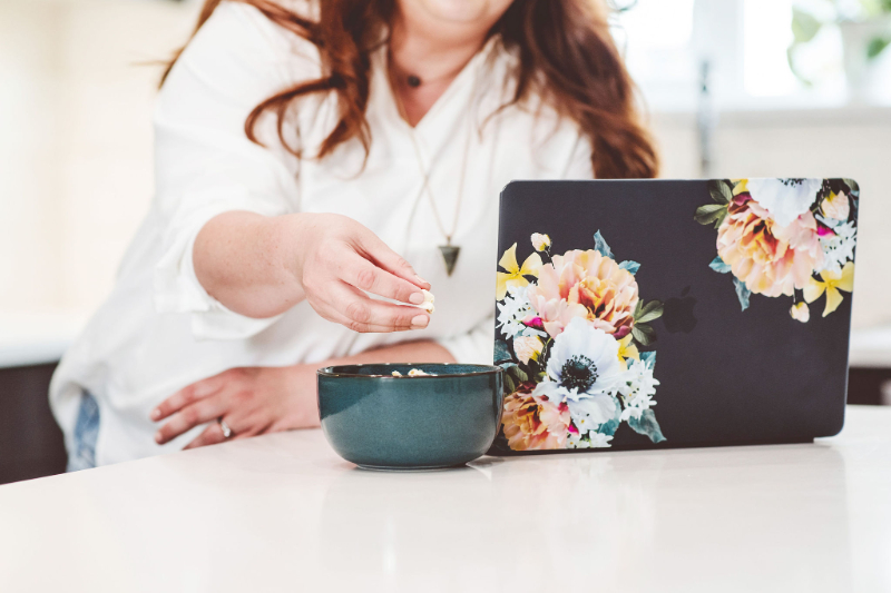 Woman snacking at laptop during clean eating cleanse, showing distracted, rule focused eating habits.