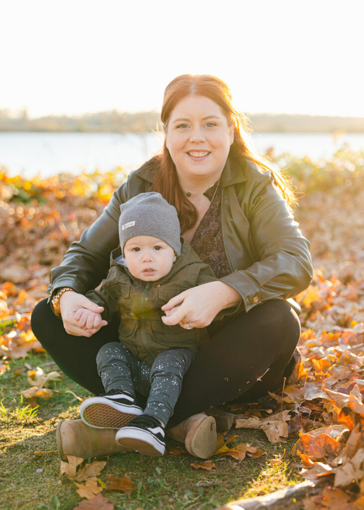 A woman sitting outdoors in fall leaves with her young child, smiling at the camera, representing a calm and balanced life while making healthy food choices.