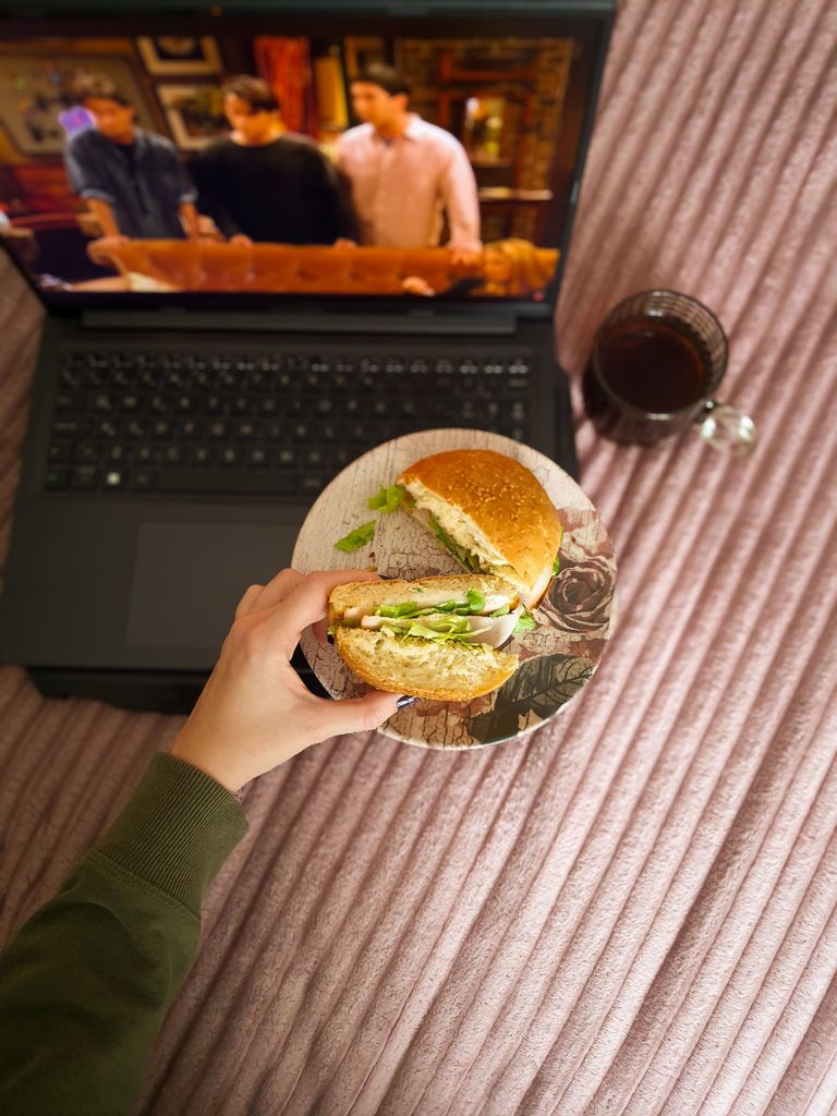 Person eating a sandwich while watching a video on a laptop, illustrating boredom eating and snacking while distracted.